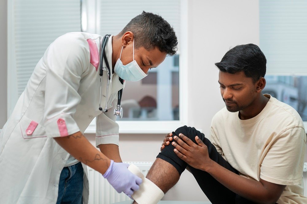 A healthcare professional in a mask wrapping a patient's injured knee with a bandage during a medical consultation.